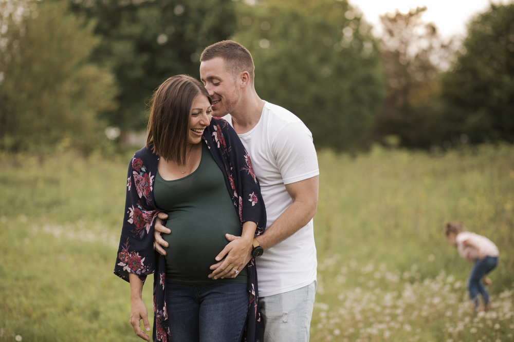 Maternity Photographer Rochester NY, Mom and Dad to be laughing  together while daughter plays in the field behind